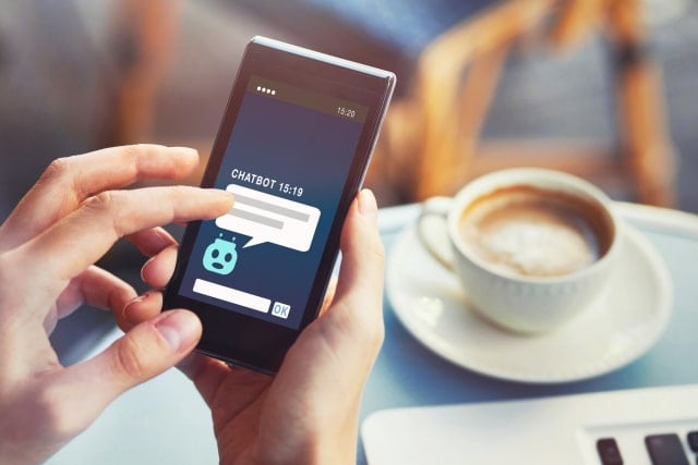 photo of someone holding a mobile phone and using an online chatbot service while they are sitting at a cafe table with a cup of coffee