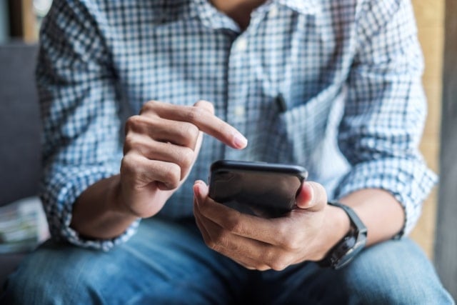 photo of a man's arms and torso, he's sitting down holding a mobile phone in one hand and using it with his other hand