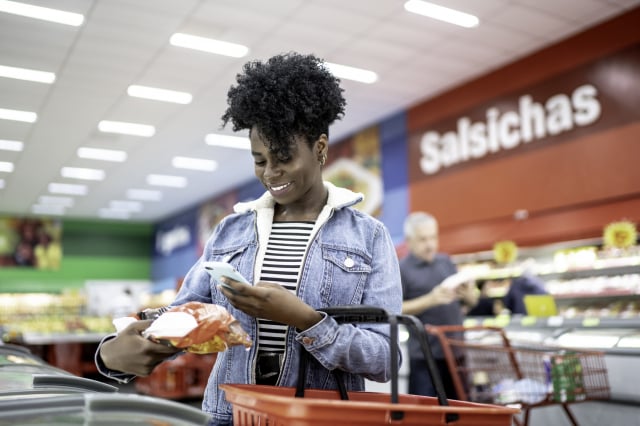 smiling young woman holding product in supermarket and searching on the phone