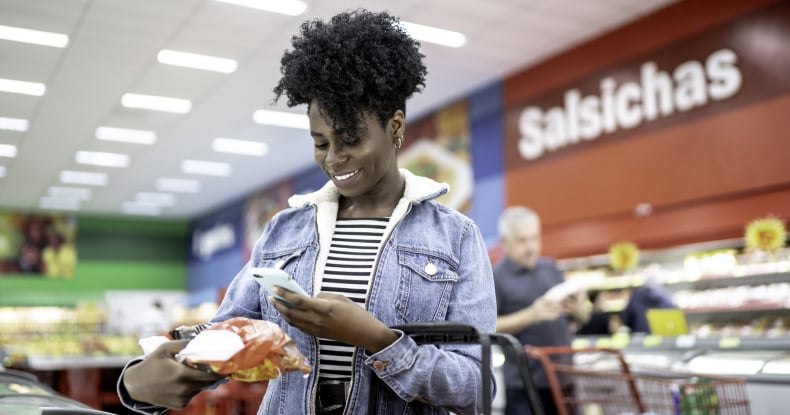 smiling young woman holding product in supermarket and searching on the phone