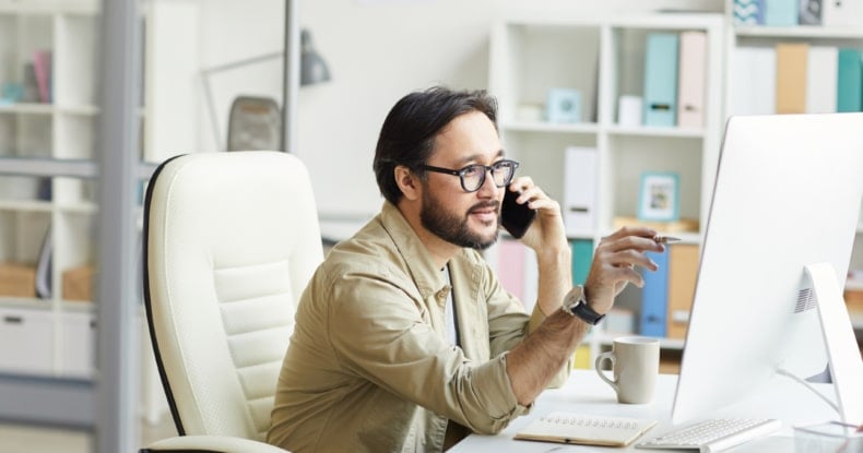 smart young asian programmer in casual shirt sitting at table with notepads and pointing at computer monitor while talking to customer by phone