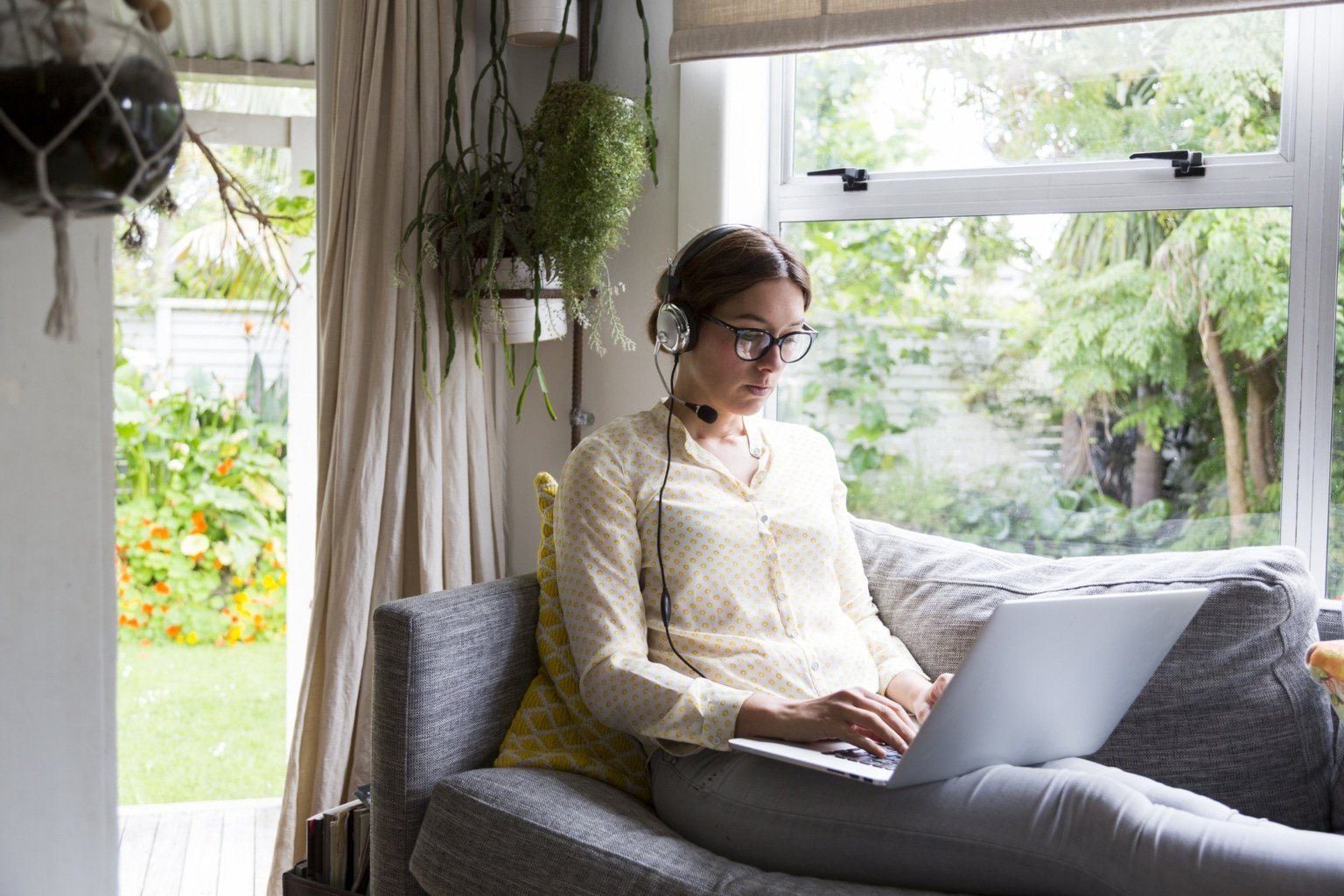 Woman working from home sitting on a sofa working on a laptop wearing a headset