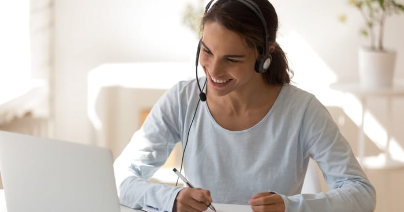 woman working at a table from home, wearing a headset and writing on a notepad while smiling at her laptop