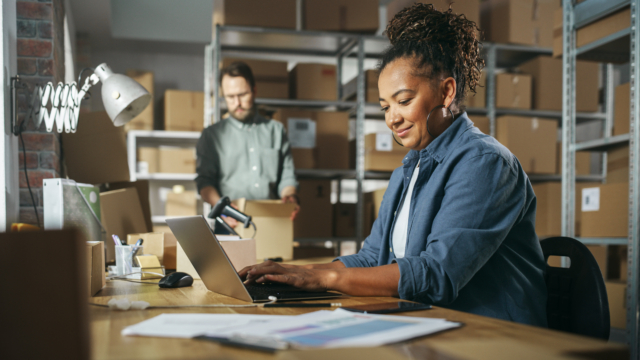 Diverse Male and Female Warehouse Inventory Managers Talking, Using Laptop Computer and Checking Retail Stock. Rows of Shelves Full of Cardboard Box Packages in the Background.-438