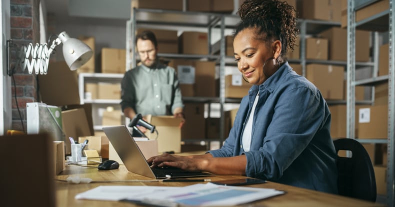 Diverse Male and Female Warehouse Inventory Managers Talking, Using Laptop Computer and Checking Retail Stock. Rows of Shelves Full of Cardboard Box Packages in the Background.-438