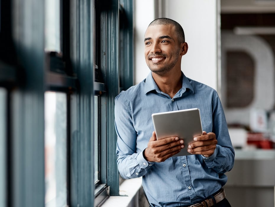 Young business person in a hybrid work schedule using a digital tablet while standing at a window in an office