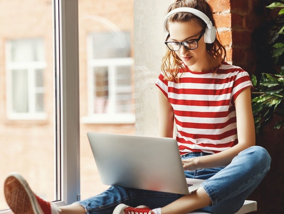 Student with headset sitting in a window typing on laptop for remote learning