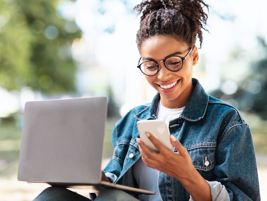 A student with glasses smiling and looking at her phone with a laptop in her lap