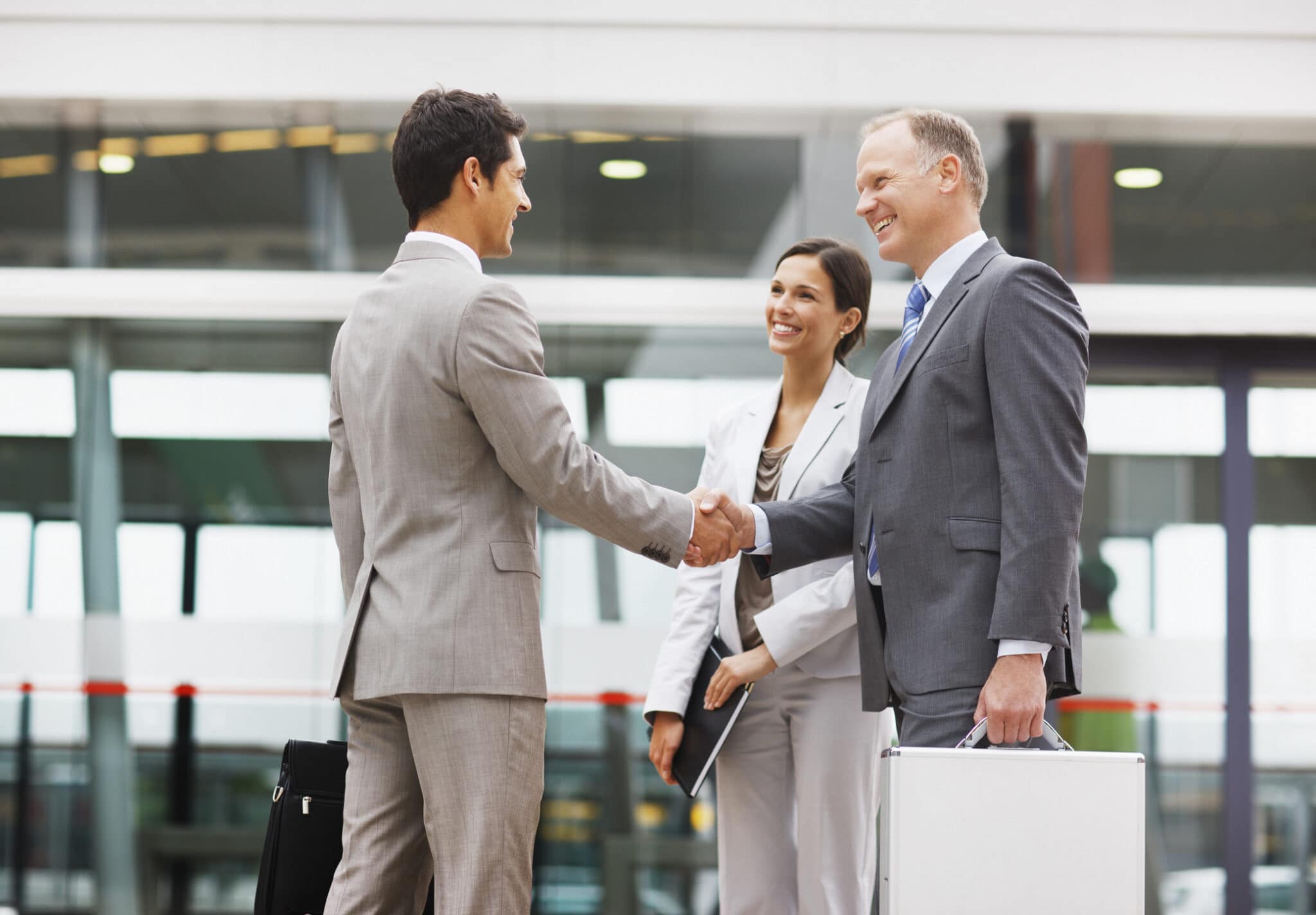 2 businessmen and 1 business woman smiling and shaking hands outside of an office