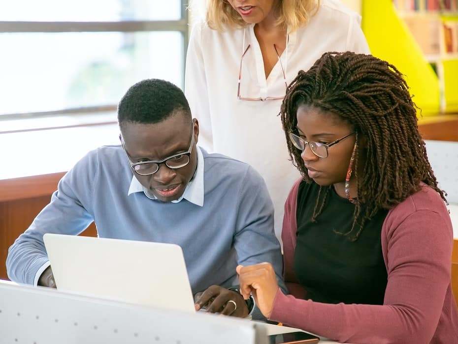 Teacher stands behind two students sitting at a table and looking at a laptop