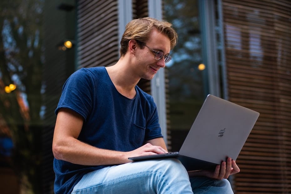 A prospective student sitting in front of an educational building on its steps using a laptop