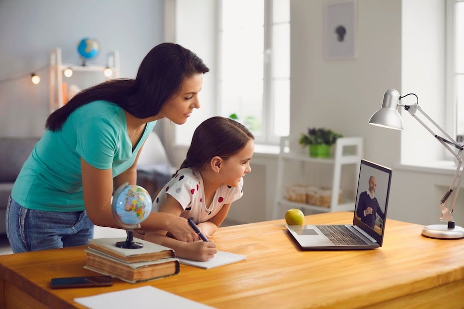 A mother leans over her daughter's shoulder in front of a laptop to help with education